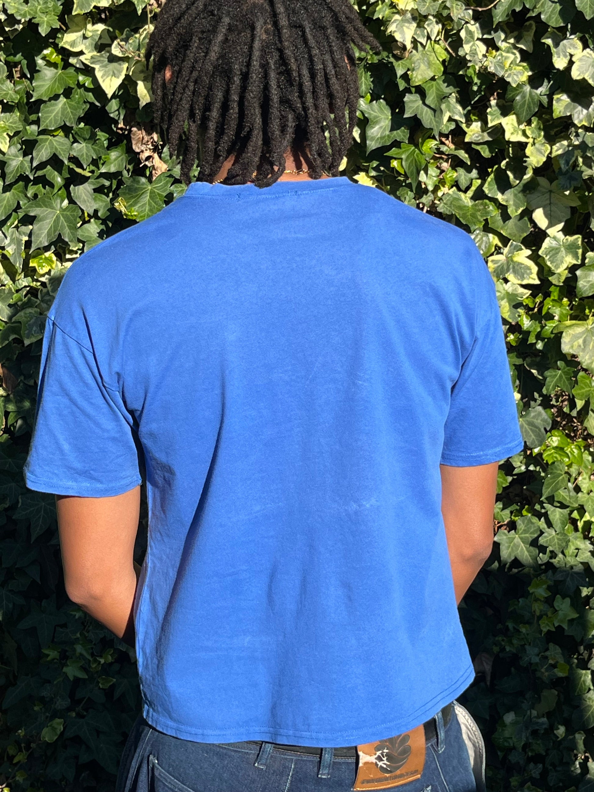 Person wearing a blue t-shirt standing against a green leafy background.