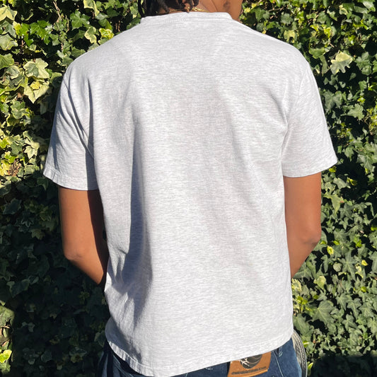 Person wearing a light grey shirt standing among green foliage